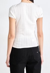 Biała bluzka z krótkim rękawem, o ribbowanej fakturze i koronkowym wykończeniu, z dopasowanym krojem i skomplikowanym wzorem pionowych pasków. Czarne spodnie z tylnymi kieszeniami.