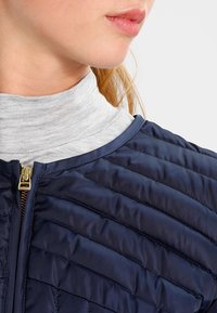 Marine pufferjack met gequiltde textuur, voorzien van een gouden rits en ronde halslijn, gelaagd over een lichtgrijze coltrui.