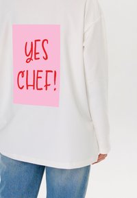 Witte long-sleeve shirt met een grote roze rechthoek op de achterkant waarin "YES CHEF!" in vetrode letters is gedrukt. Casual pasvorm, zachte stof.