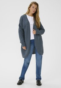 Femme debout portant un long cardigan en tricot gris foncé, une chemise blanche à boutons, un jean bleu et des mocassins noirs, sur un fond uni.