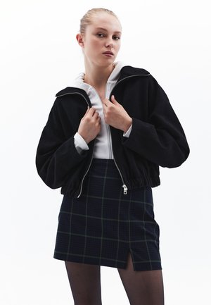 Giacca nera corta con colletto alto, abbinata a una maglia bianca con zip e una mini gonna a scacchi scuri con spacco laterale.