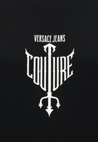 Λευκό κείμενο "VERSACE JEANS COUTURE" με στυλιζαρισμένο σχέδιο τρίαινας σε μαύρο ύφασμα.