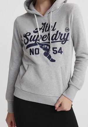 Grijze hoodie met "Atel Superdry No 54" en vleugelvormig logo in marineblauw, gedragen door een persoon waarvan de handen gedeeltelijk zichtbaar zijn.