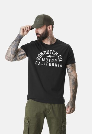 T-shirt en coton noir avec le texte graphique blanc "VON DUTCH CO. MOTOR CALIFORNIA". Modèle portant une casquette verte, mettant en avant des tatouages sur les bras et un pantalon cargo.