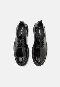 Zapatos de charol negro con punta redondeada, diseño con cordones y suela texturizada. Presenta una pequeña lengüeta en el talón. Logotipo visible en el interior.