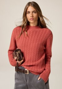 Suéter de punto trenzado color óxido con cuello alto y mangas acanaladas, combinado con un bolso de mano marrón oscuro y pantalones de pana grises.