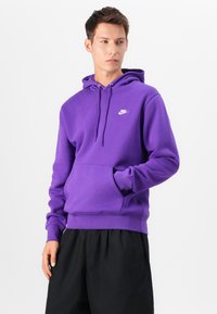 Hanorac violet din amestec de bumbac, cu buzunar frontal, glugă cu șnur și logo alb Nike pe piept. Port cu pantaloni scurți negri.