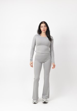 Ung kvinde med langt mørkt hår iført en tætsiddende lysegrå langærmet top og matchende udvidede bukser med sneakers, stående ret op.