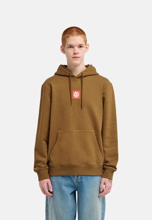 Personne aux cheveux courts et roux porte un sweat à capuche marron avec un écusson logo rouge et un jean bleu, debout devant un fond uni clair.