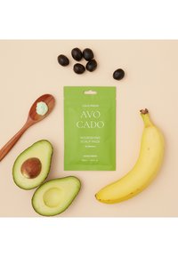 Grön påse med etiketten "AVO CADO Nourishing Scalp Pack" bredvid en träsked, en halverad avokado, en banan och svarta olivliknande frukter.
