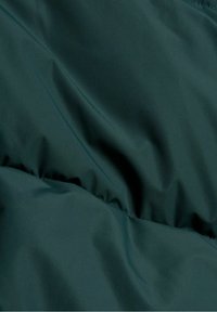 Ciemnozielona tkanina o gładkiej fakturze, z delikatnymi fałdami i błyszczącym wykończeniem, charakterystyczna dla materiału odzieży wierzchniej. Brak wzorów i elementów metalowych.