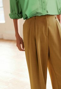 Kaki geplooide broeken met een op maat gemaakte pasvorm, gemaakt van lichtgewicht stof; heeft zijzakken en een soepele textuur. Kreukelig groen shirt zichtbaar.