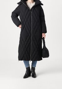 Lange zwarte doorgestikte donsjas met een rits, capuchon en diagonaal patroon. Gepaard met blauwe jeans en zwarte enkellaarzen, met een gestructureerde zwarte tas.