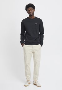 Felpa nera a maniche lunghe con collo rotondo e logo ricamato sul petto, abbinata a pantaloni tapered di un leggero beige e sneakers bianche.