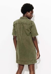 Robe en denim verte courte à manches courtes, avec un col boutonné, un détail de pli au dos et des poches latérales, présentant une texture de tissu douce.
