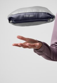 Sac de rangement gris avec une fermeture éclair marine, de forme ovale, texture lisse, et étiquette "STOW AWAY". Il est tenu au-dessus d'une main ouverte sur un fond neutre.