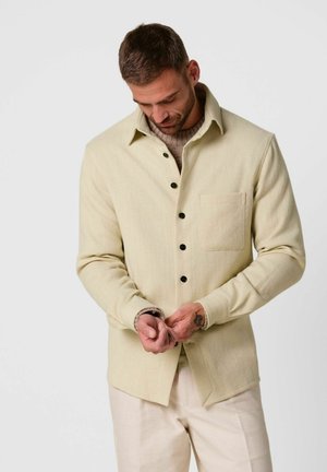 Uomo che allaccia il polsino di una giacca camicia beige sopra un maglione di maglia, indossa pantaloni chiari, in piedi contro uno sfondo semplice.