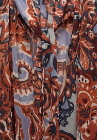 Tissu transparent avec un motif floral dans des tons rouille, marine et bleu clair. Présente une forme fluide avec des plis horizontaux et un détail à nouer.