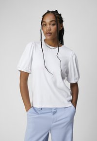 Haut blanc côtelé avec des manches bouffantes, assorti à un pantalon sur mesure bleu clair. Le mannequin porte un collier en chaîne et a les cheveux tressés.