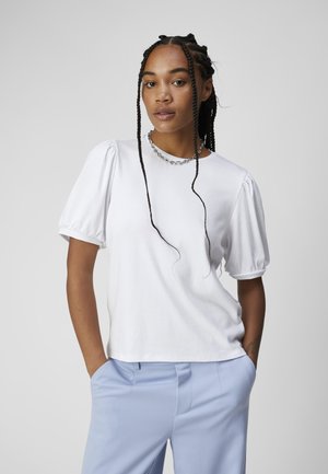 Haut blanc côtelé avec des manches bouffantes, assorti à un pantalon sur mesure bleu clair. Le mannequin porte un collier en chaîne et a les cheveux tressés.