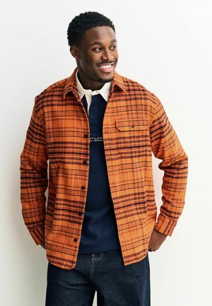 Oranges karierter Hemd aus Stoff mit schwarzen Akzenten, mit Knopfleiste, Kragen und einer Brusttasche, über einem blauen Sweatshirt getragen.