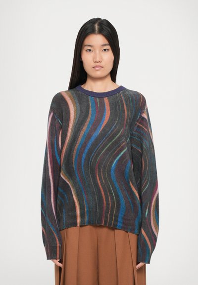 Donna che indossa un maglione scuro con strisce ondulate multicolori e una gonna marrone, in piedi contro uno sfondo bianco semplice.