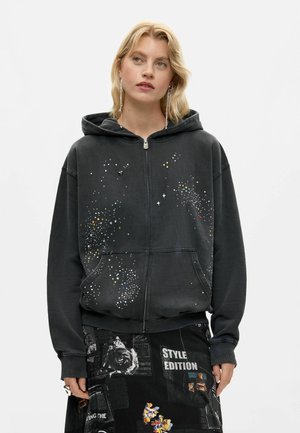 Blonde woman wearing a black zip-up hoodie with colorful speckles and a black skirt with mixed graphic prints and text.
