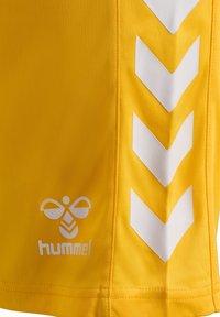Geel sportshorts gemaakt van lichtgewicht stof. Heeft witte chevrons langs de zijkanten en een logo met een bij en de tekst "hummel."