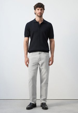 Homme debout portant un polo en maille noire à manches courtes, un pantalon gris clair et des mocassins noirs sur un fond clair uni.