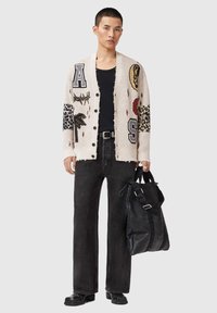 Πλεκτή ζακέτα με διάφορα γραφικά, εκρού βάση, μαύρα κουμπιά, φορεμένη πάνω από μαύρο πουκάμισο. Συνδυασμένη με μαύρο τζιν και μεγάλη μαύρη τσάντα.