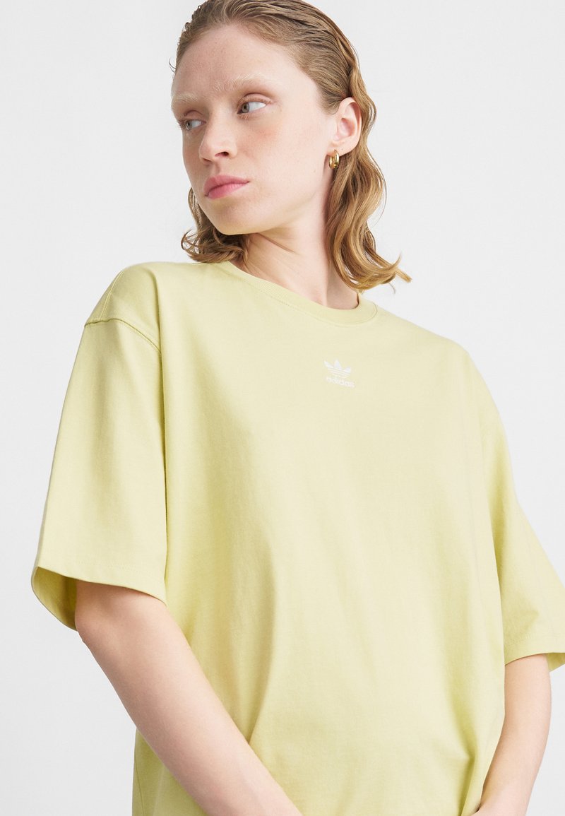 Žena s krátkými mokrými vlasy, která nosí zlaté kruhové náušnice a světle žluté oversized tričko Adidas, na bílém pozadí.
