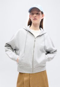 Νεαρό άτομο που φοράει ανοιχτό γκρι φούτερ με φερμουάρ, καρό καφέ φούστα, λευκό πουκάμισο και ανοιχτό μπλε καπέλο, στέκεται με τα χέρια στις τσέπες.