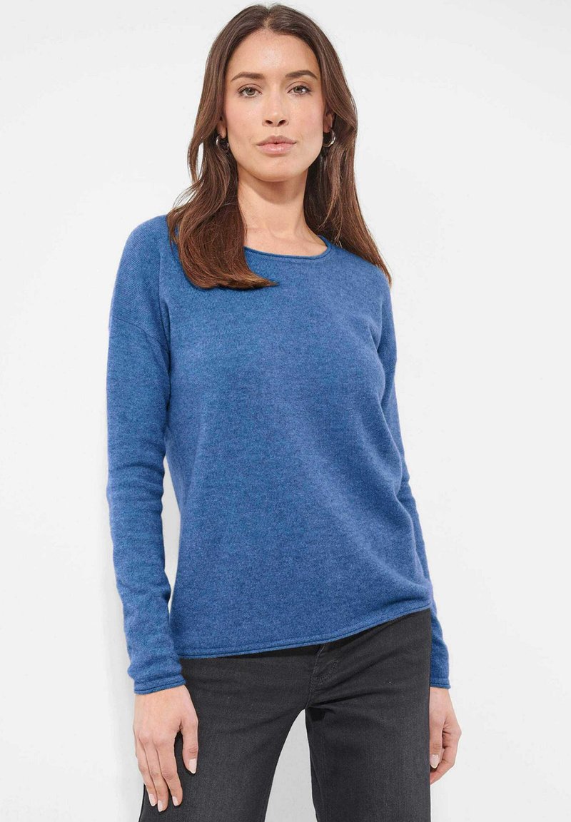 Frau mit langen braunen Haaren, die einen blauen langärmligen Pullover und schwarze Hosen trägt, steht vor einem schlichten weißen Hintergrund.