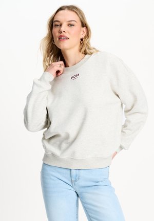 Vrouw met blond haar die een lichtbeige sweatshirt met het logo "POM" en lichtblauwe spijkerbroek draagt, poseert met één hand bij haar nek.