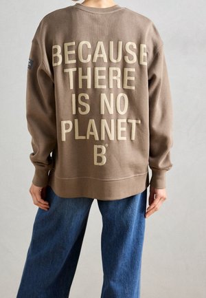 Osoba nosząca brązową bluzę z dużym beżowym napisem „BECAUSE THERE IS NO PLANET B” na plecach, w połączeniu z niebieskimi dżinsami.