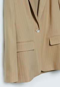 Ljusbrun kavaj med vertikala vita ränder, enkel knappstängning, två framfickor och en texturerad tygyta.