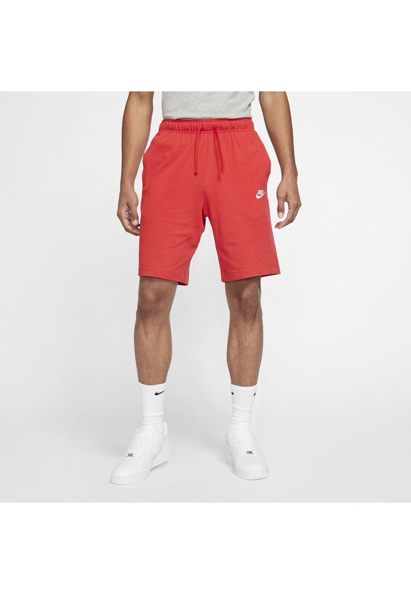 Homme portant un short rouge Nike, des baskets blanches et des chaussettes blanches Nike, debout devant un fond blanc uni.