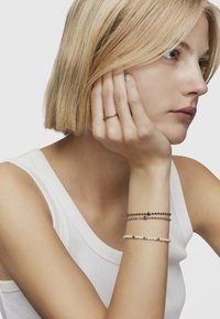 Anillo de oro en una mano que sostiene la cara; tres pulseras: cuentas negras, piedras preciosas de varios colores y perlas blancas con acentos de oro.
