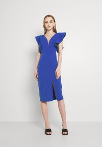 Vestido azul justo com mangas em babados, decote em V profundo e uma fenda frontal. Textura suave e design até ao joelho. Sapatos de salto aberto pretos.