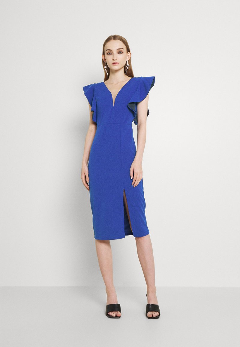 Vestido azul justo com mangas em babados, decote em V profundo e uma fenda frontal. Textura suave e design até ao joelho. Sapatos de salto aberto pretos.