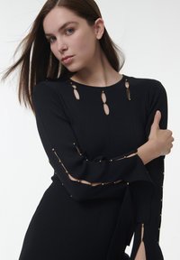 Schwarzes, geribtes Langarmkleid mit goldenen Akzenten, Schlüssel-ausschnitten am Ausschnitt und an den Ärmeln sowie einem fließenden Saum.