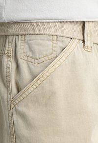 Un pantalon en coton beige présente une ceinture texturée beige, des poches frontales inclinées et des détails de couture légers le long des coutures et des poches.