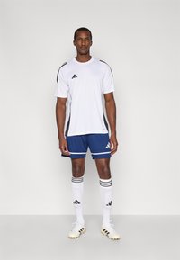 Weißes Sport-T-Shirt mit schwarz-weißen Streifen, kombiniert mit blauen Shorts. Dazu weiße Socken mit schwarzen Streifen und hellfarbene Fußballschuhe.