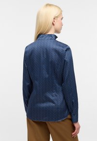 Camicia a maniche lunghe in raso blu con un motivo geometrico, caratterizzata da un colletto alto e una silhouette aderente. Tessuto morbido con polsini abbottonati.
