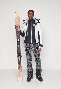 Sífelszerelés, amely egy fehér, pufi kabátot, tengerészkék alapréteget, absztrakt mintájú nadrágot és fekete síbakancsokat tartalmaz. Egy rózsaszín és barna síléccel a kezében.