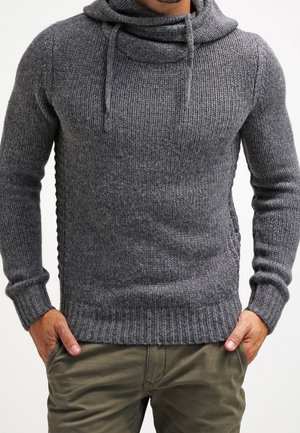 Homme portant un pull gris en maille avec un col bénitier et des cordons, associé à un pantalon vert olive, mains dans les poches.
