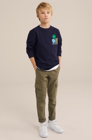 Námornícky sveter s grafickým dizajnom v zelenej a modrej farbe, v kombinácii s olivovo zelenými cargo nohavicami so bočnými vreckami. Biely tenisky dotvárajú outfit.