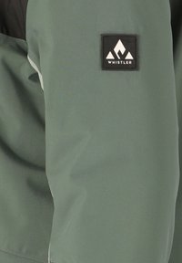 Groene jas van een gladde, matte stof, met een zwart logo-label met bergafbeelding en contrasterende horizontale en verticale naden.