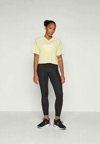 Gaiši dzeltena īsa T-krekls ar baltu logotipu, kombinēts ar melnām legingām un melniem sporta apaviem ar rozā akcentiem. Īsas piedurknes un brīvs piegriezums.