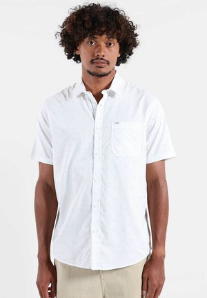 Homme aux cheveux bouclés portant une chemise blanche à manches courtes avec une poche poitrine, debout devant un fond clair uni.